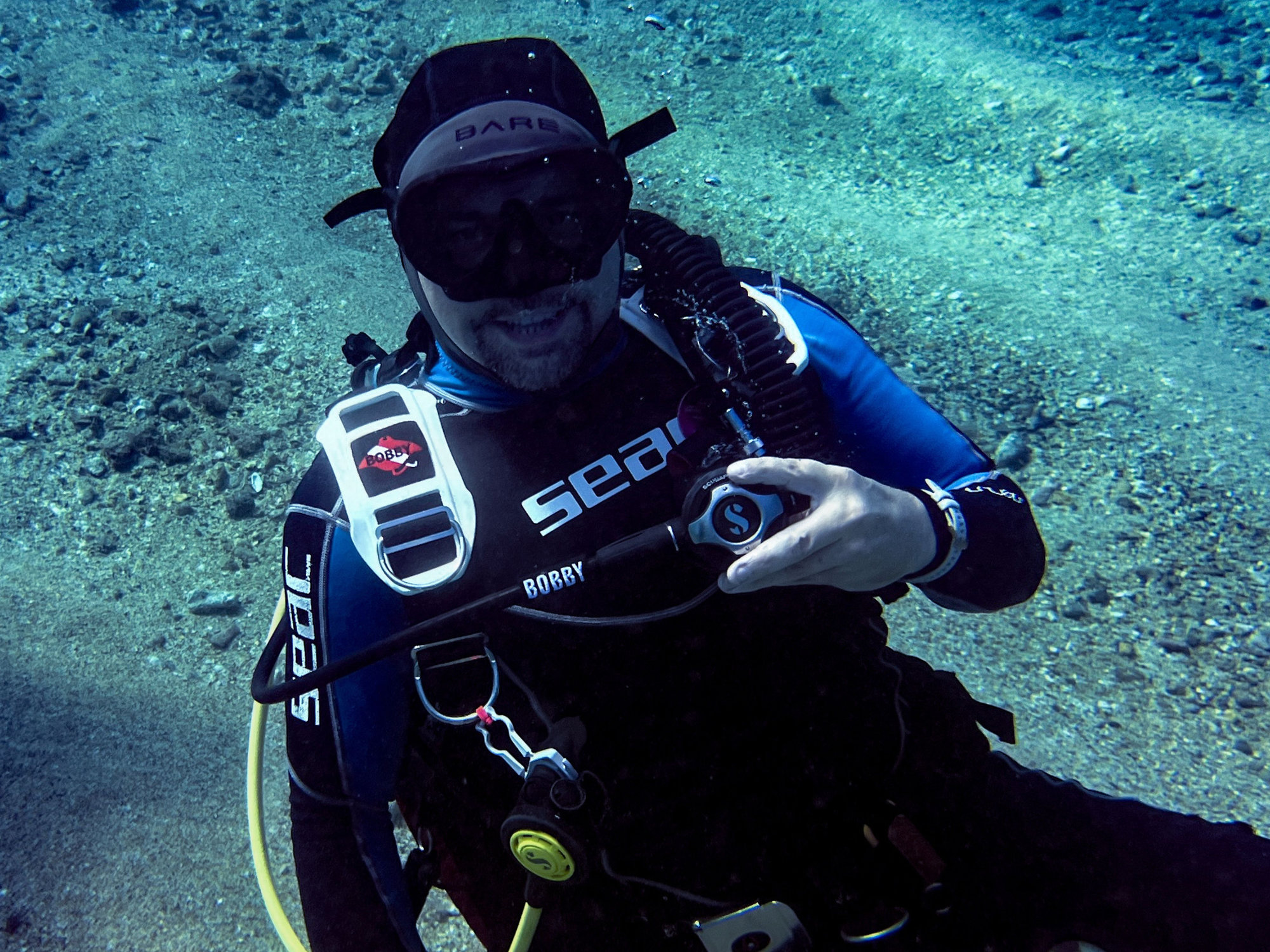 Scuba diver underwater wearing a Rinn Stitches BCD Tag featuring a Dive Flag Manta Ray in Red and White with the name...